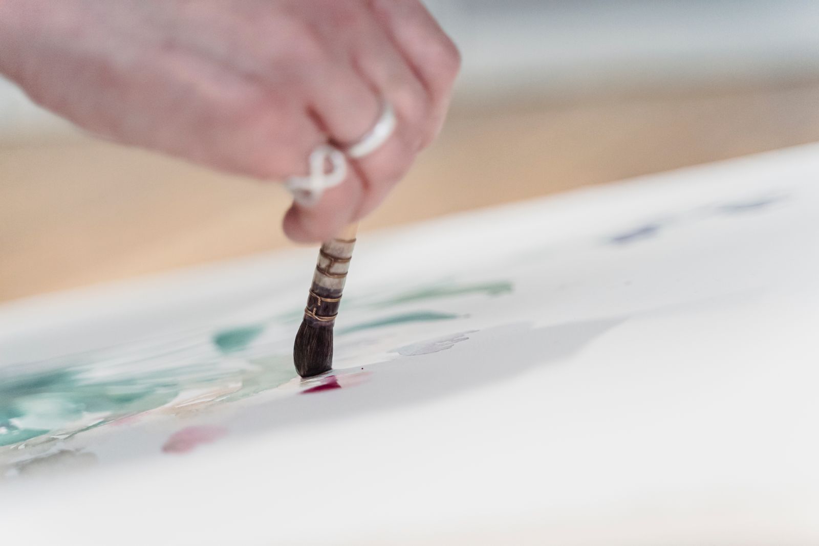 A hand guiding a watercolor brush across pale green and rose washes on paper